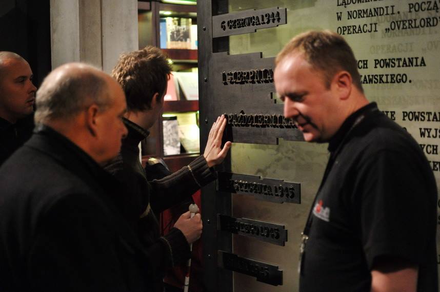 Na zdjęciu mężczyzna dotyka napisów z wypukłych liter umieszczonych na ścianie. Są to daty wydarzeń z Powstania Warszawskiego.