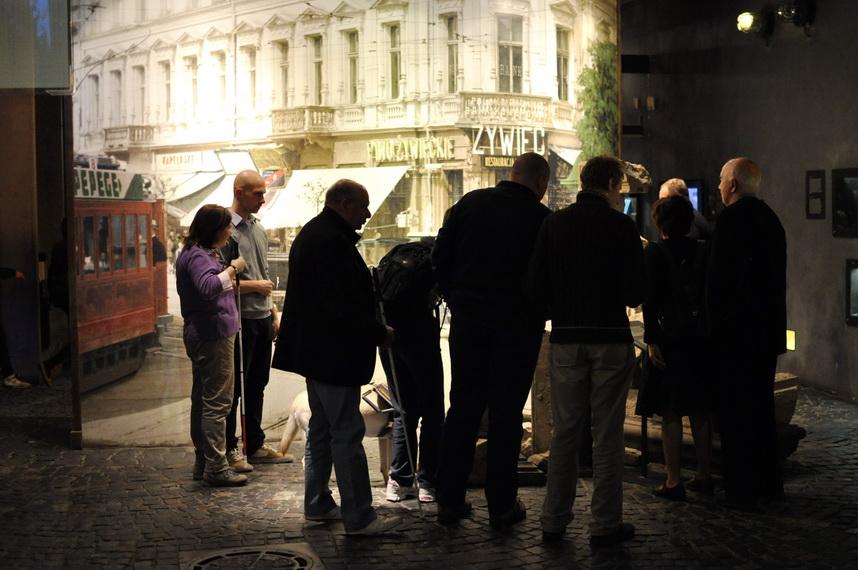Na zdjęciu grupa osób stoi przed ekranem. Na ekranie wyświetlone jest przedwojenne zdjęcie okazałej, narożnej kamienicy z neonem „Żywiec”